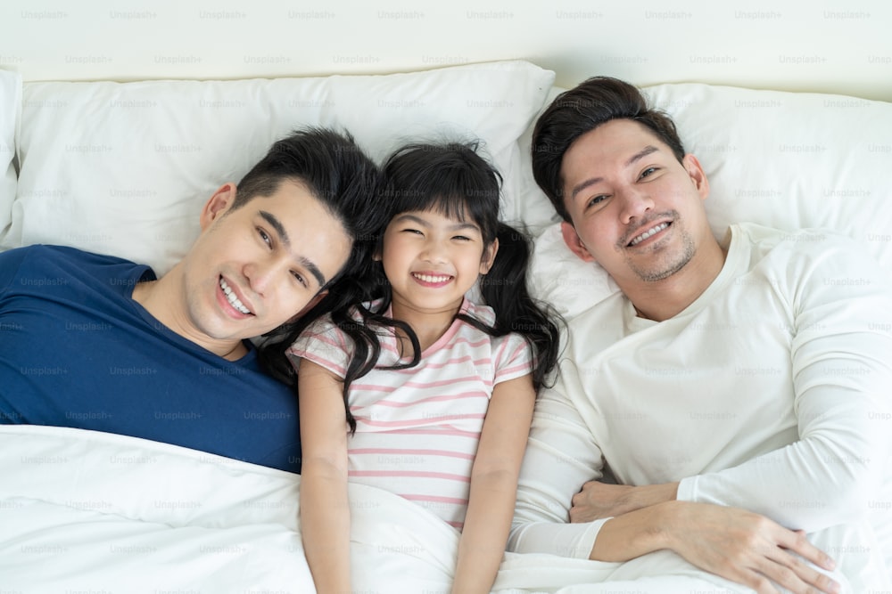 Portrait of handsome man gay family with young kid daughter in bedroom. Attractive romantic male lgbt couple lying down on bed with little adorable girl child in morning and looking at camera in house