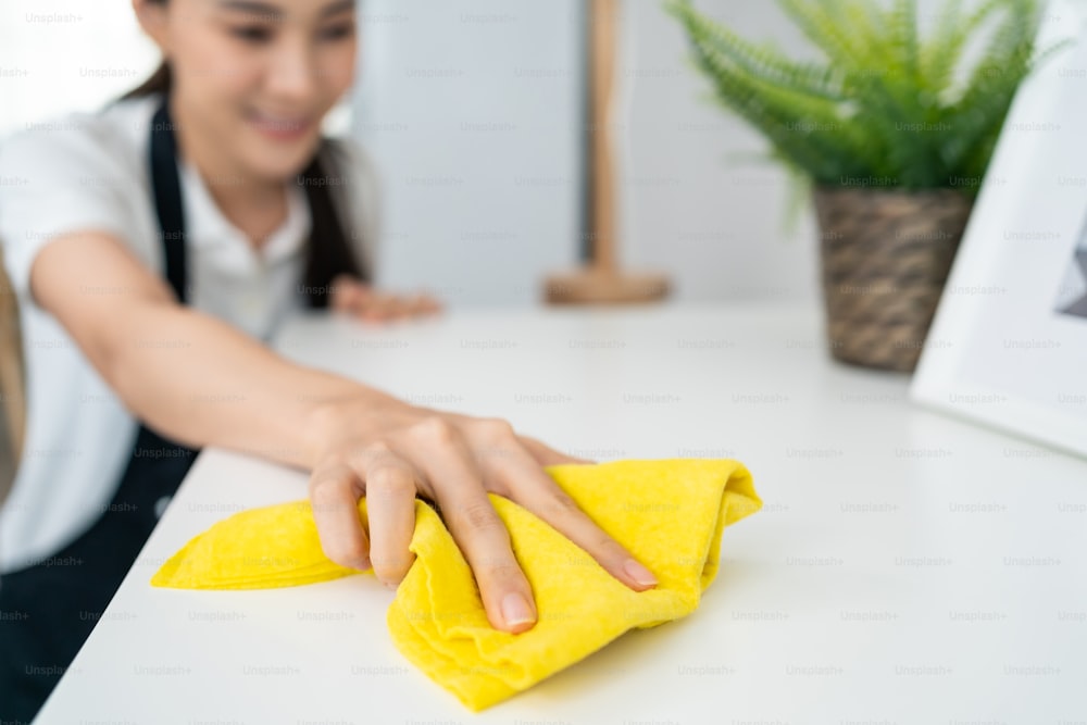 Asiatische Reinigungsdienstmitarbeiterin putzt im Wohnzimmer zu Hause. Schöne Mädchen Hausfrau Haushälterin Reinigungskraft fühlen sich glücklich und wischen unordentlichen schmutzigen Arbeitstisch für Housekeeping Hausarbeit oder Hausarbeiten.