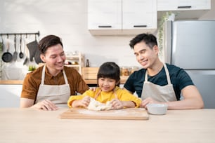 Asian attractive LGBTQ gay family teach girl kid making yeast dough. Handsome male couple take care and spend time with little adorable child bake bakery in kitchen, enjoy parenting activity at home.