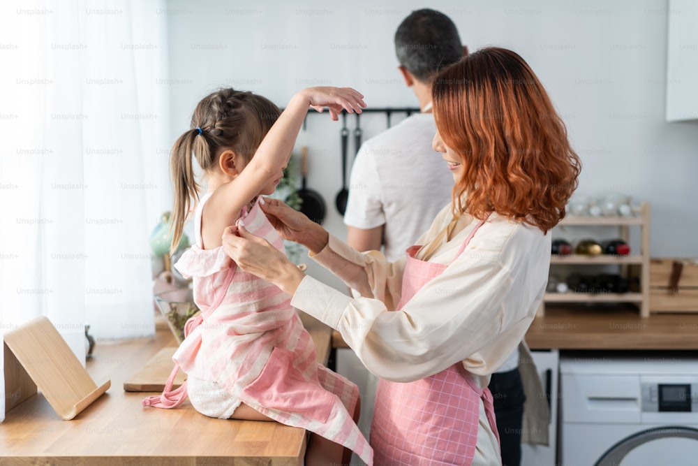Asian beautiful young mother cooking foods with baby kid in kitchen. Happy family spend time on holiday together in house, loving parent tying apron for little child daughter prepare to make breakfast