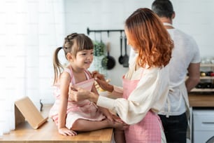 Asian beautiful young mother tying apron for baby kid before cooking. Happy family spend time on holiday together in house, loving parent make foods breakfast for little child daughter in kitchen room