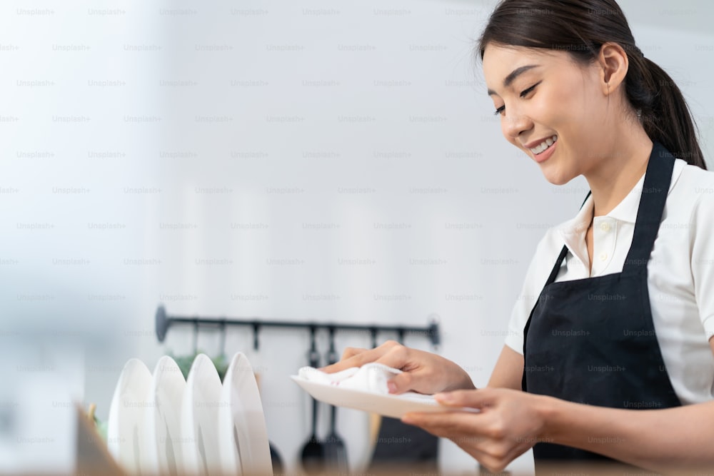 Asian active cleaning service woman worker cleaning in kitchen at home. Beautiful young girl housekeeper cleaner feel happy and wipes dishes plates after washing for housekeeping housework or chores.