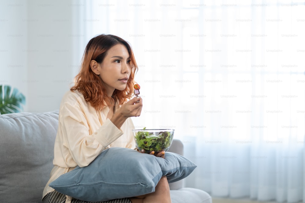 Asiatische junge glückliche attraktive Frau essen grünen Salat, während Film schauen. Schönes Mädchen fühlen sich fröhlich und genießen es, Gemüse gesunde Lebensmittel zu essen Diät und Gewicht zu verlieren für die Gesundheitsfürsorge Wellness im Haus.