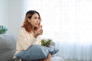 Asian young happy attractive woman eat green salad while watch movie. Beautiful girl feel joyful and enjoy eating vegetables healthy foods to diet and lose weight for health care wellness in house.