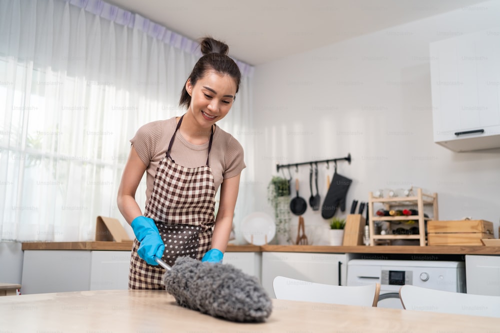 Femme de ménage asiatique qui nettoie dans la cuisine à la maison. Belle jeune femme de ménage femme de ménage porter un tablier et utiliser un plumeau essuyant la table sale pour les tâches ménagères ou les tâches ménagères.