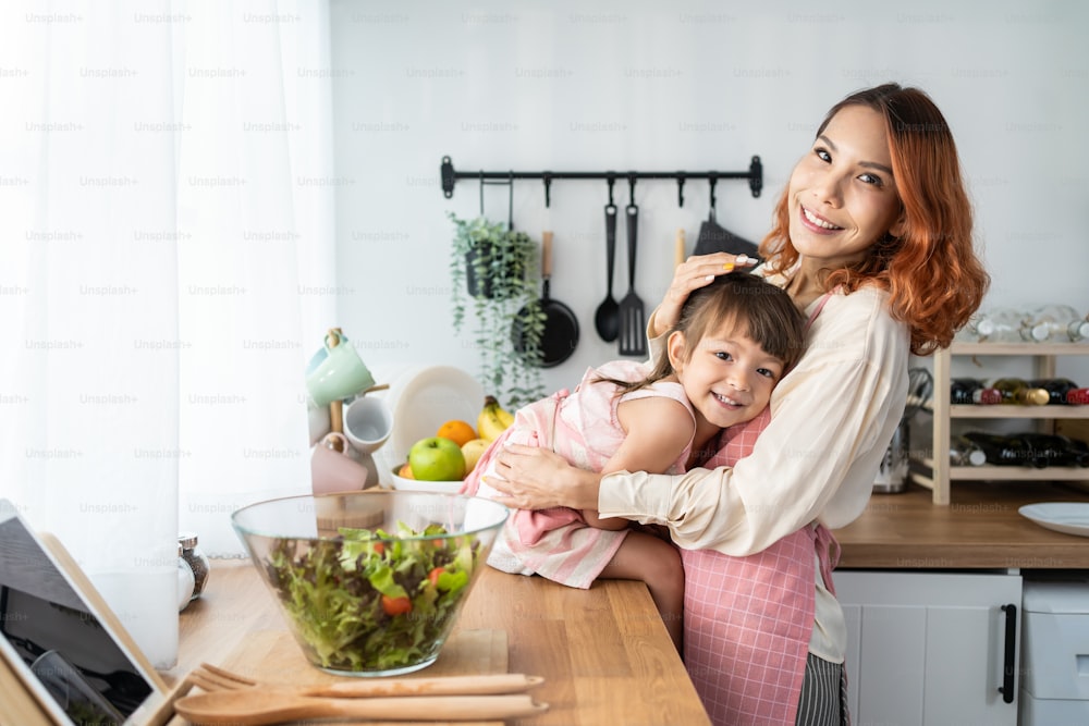 Asian happy family, Parent spend free time with kid together at home. Happy Young girl daughter sit on cooking counter with Mother in kitchen and smile, look at camera. Activity relationship in house.