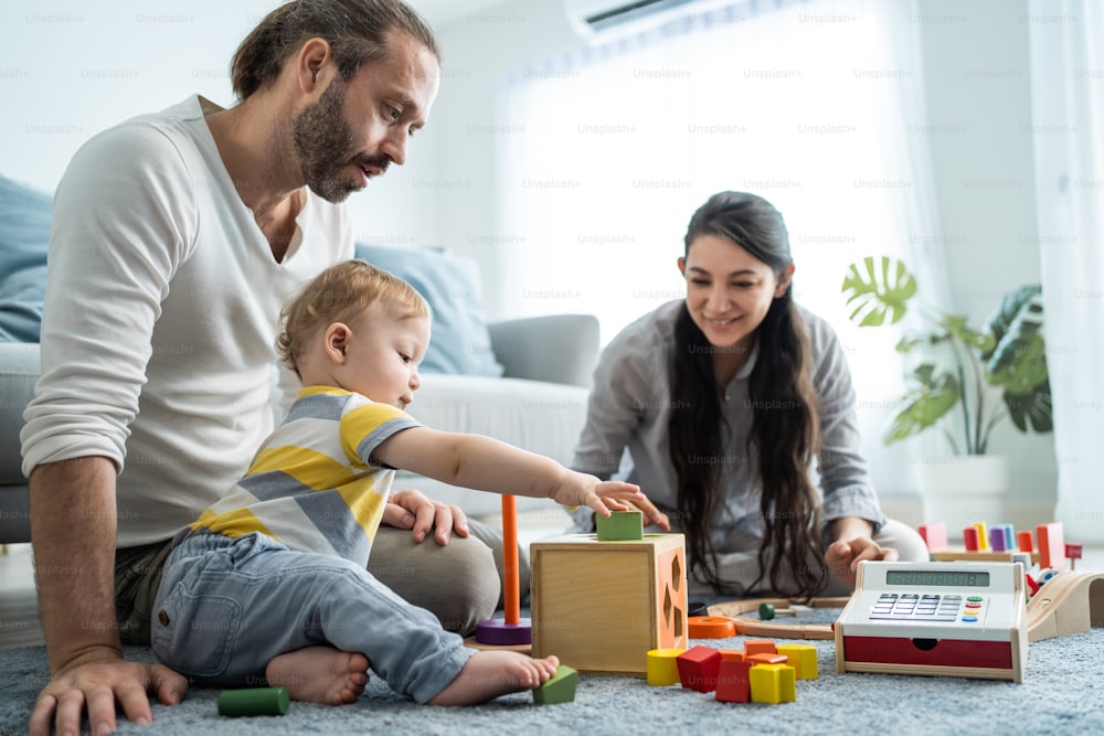 Caucasian loving parent looking at baby toddler playing in living room. Attractive couple mother and father watch young little infant son child's development. Activity relationship at home in house.