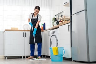 自宅の台所で掃除をしているアジアのアクティブクリーニングサービス女性労働者。美しい若い女の子の家政婦の掃除人は幸せを感じ、家事や家事のために床でモップをかけています。