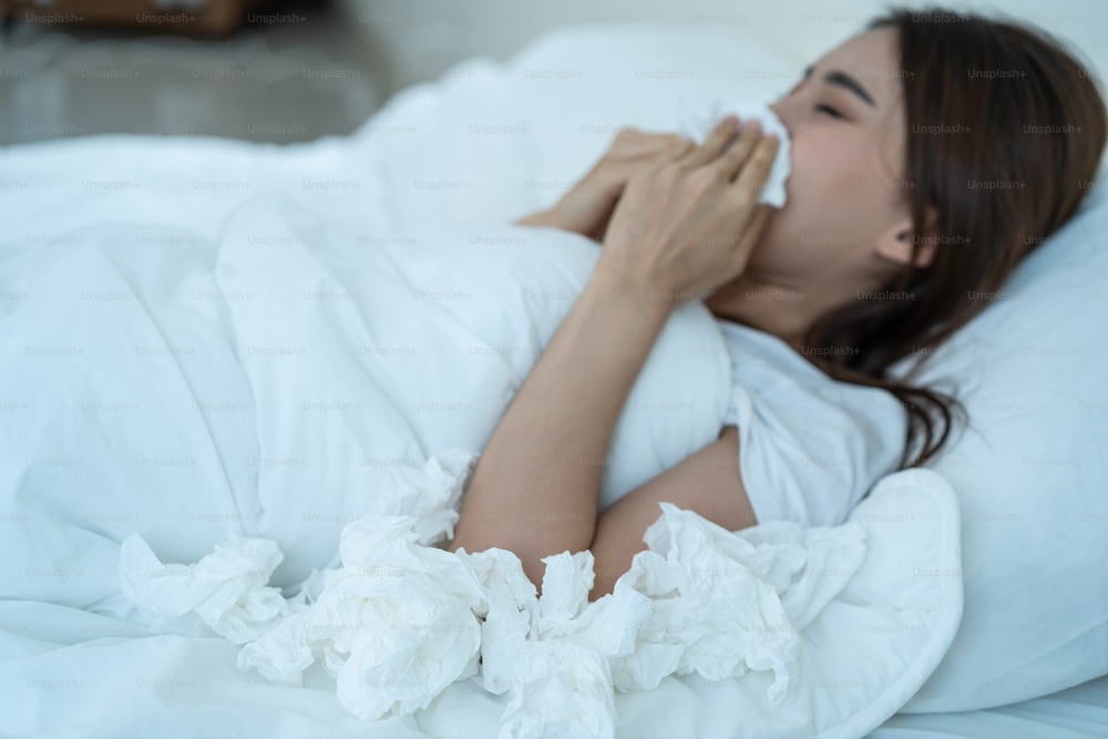 Asian sick girl in pajamas wake up from sleep at night sneezing on bed. Attractive young woman feeling bad and suffer from allergy, put tissue cover her nose while sneezes during bedtime in house.