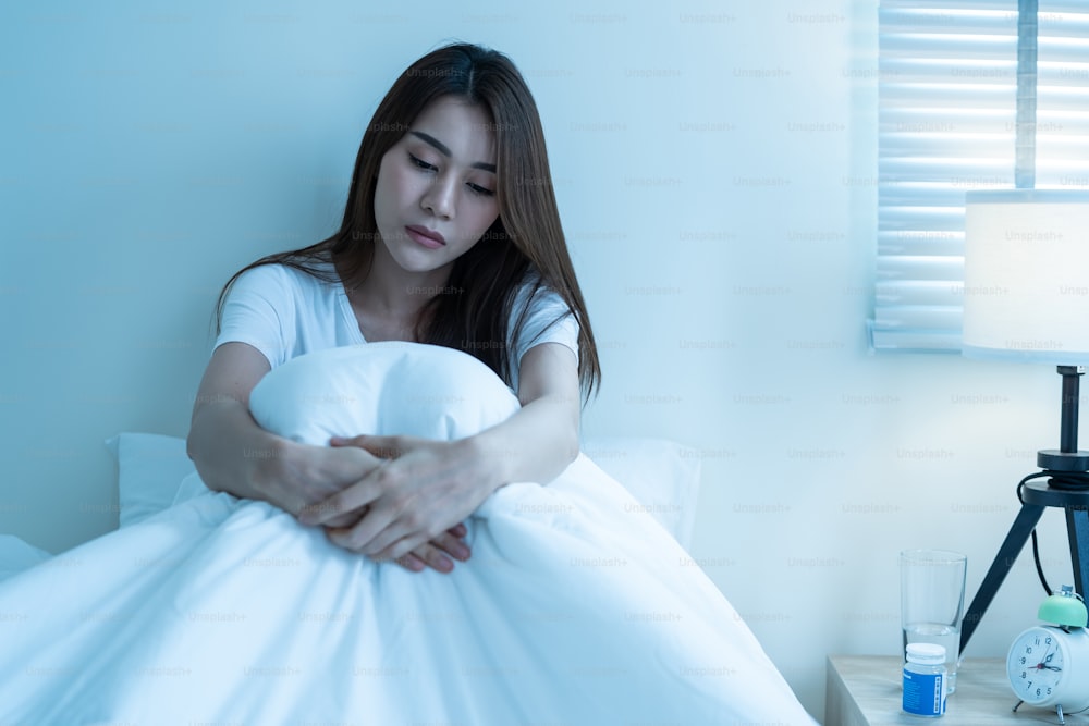 Asian beautiful upset depressed girl sitting alone on bed in bedroom. Attractive unhappy young woman feel sad lonely and upset with life problem and trying to hurt herself in dark night room at home.
