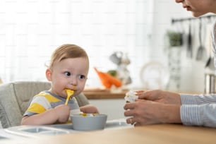 Caucasian beautiful mother take care of baby boy toddler in kitchen. Happy family, Attractive loving mom cook and feeding healthy foods to little kid son infant while sit on chair for lunch in house.