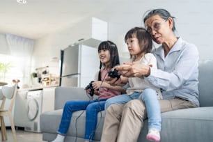 Asian happy family stay home, grandmother play game with little girl. Loving senior elder older woman play video games in front of television with young children granddaughter in living room together.