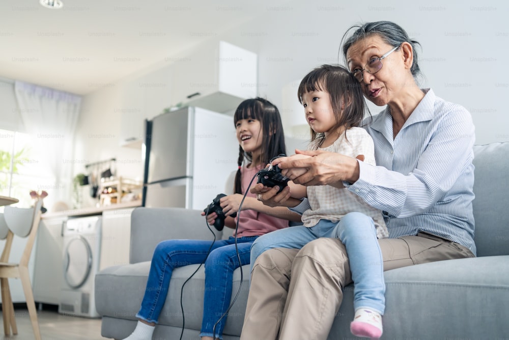 Asian happy family stay home, grandmother play game with little girl. Loving senior elder older woman play video games in front of television with young children granddaughter in living room together.