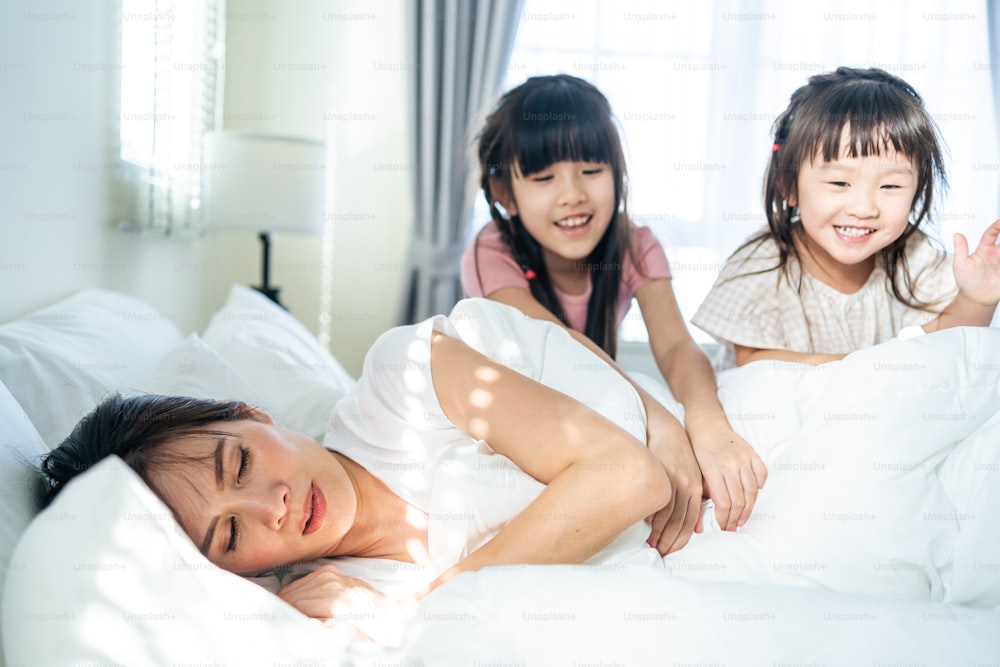 Asian happy children jumping on bed to waking up mother in the morning. Young little girl sibling daughters enjoy play with loving mom in bedroom together with happiness. Activity relationship concept