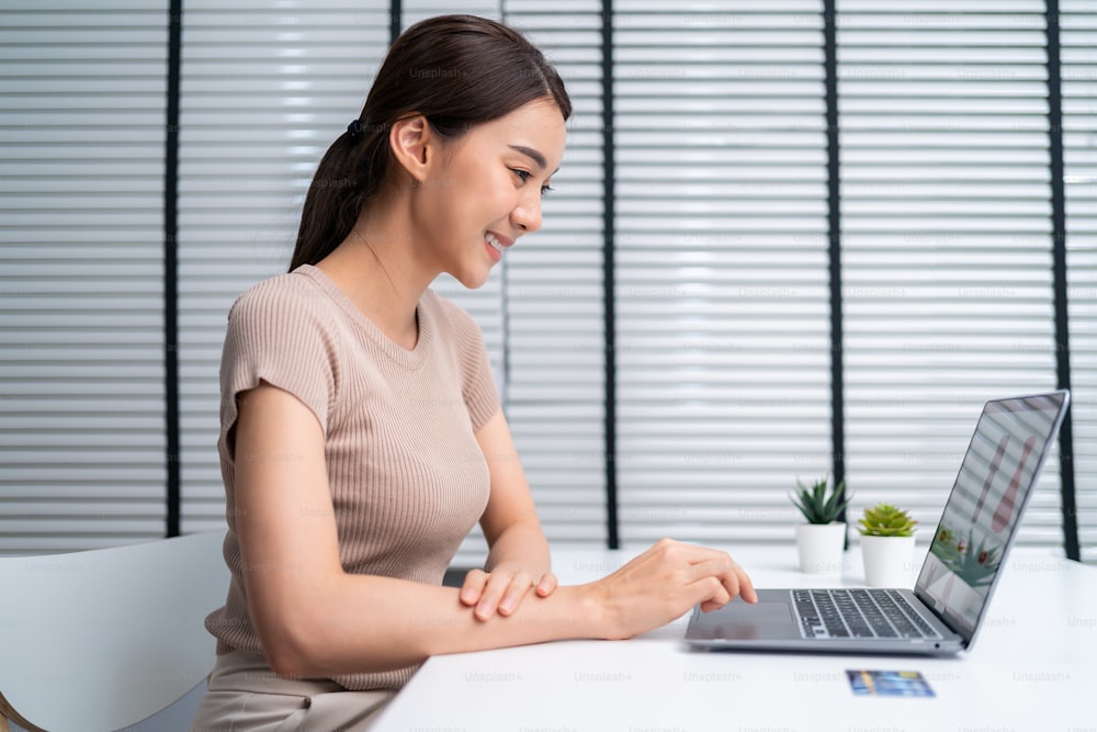 Una mujer joven asiática usa una computadora portátil para comprar productos que se venden en línea. Atractiva hermosa muchacha sosteniendo la tarjeta de crédito en las manos comprando y comprando productos en el sitio web en el teléfono inteligente. Concepto de comercio electrónico.