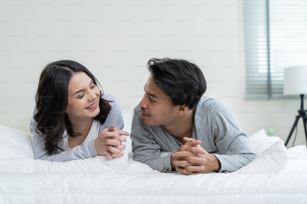 Portrait of Asian new marriage couple lying on bed and look each other. Attractive beautiful young man and woman in pajamas enjoy early morning activity in bedroom at home. Family relationship concept