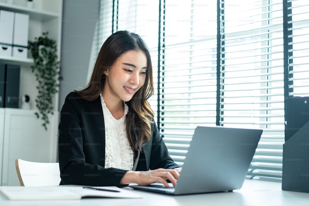 Asian happy beautiful businesswoman in formal suit work in workplace. Attractive female employee office worker smile, use laptop computer for discuss with co worker and take note on paper in company.