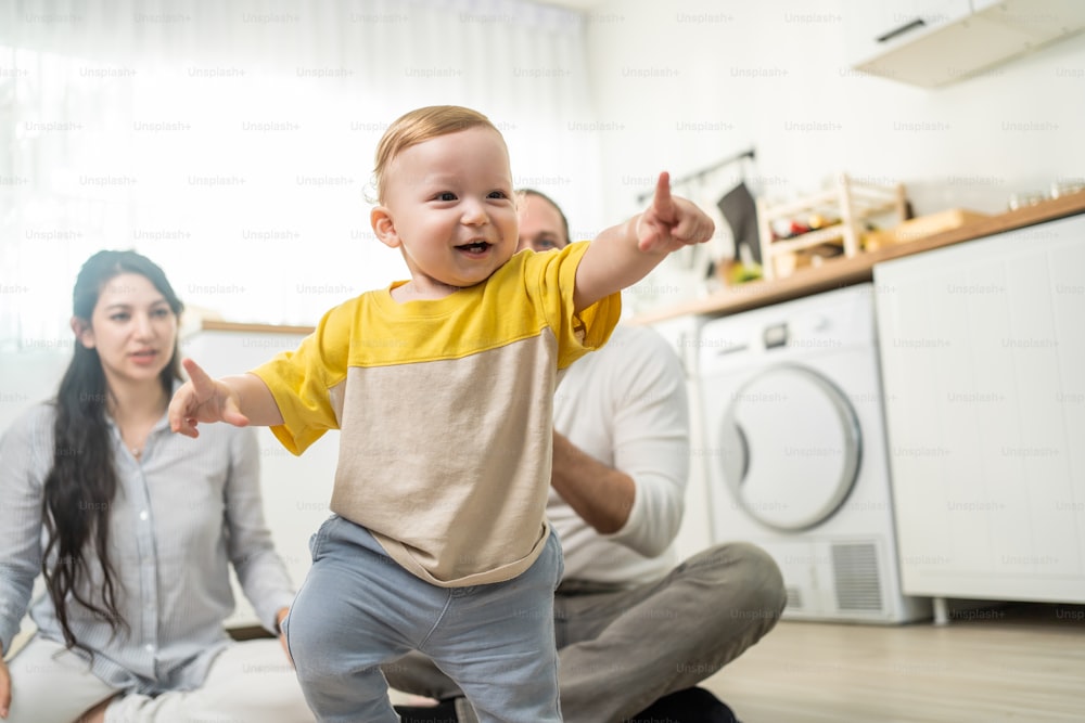 Caucasian baby boy child learn to walk with parents support in house. Happy family, mother and father helping young toddler son taking first step walk on floor to develop skill in living room at home.