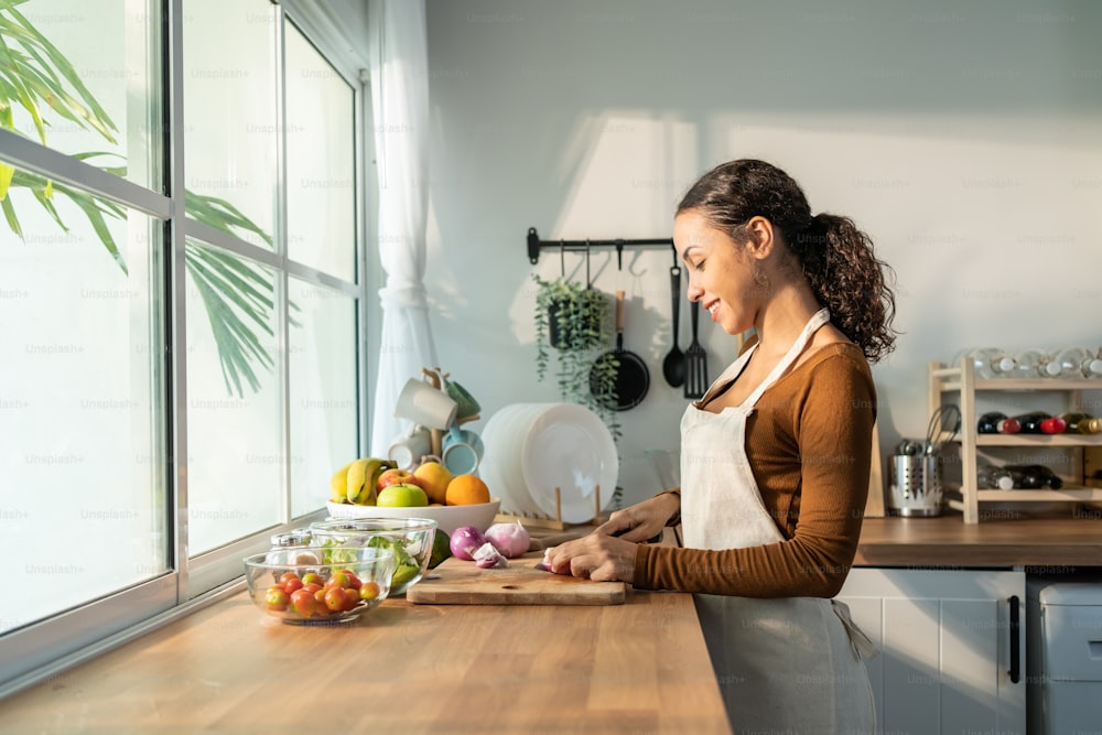 Latino attractive woman wear apron cook green salad in kitchen at home. Young beautiful girl feeling happy and enjoy eating vegetables healthy foods to diet and lose weight for health care in house.