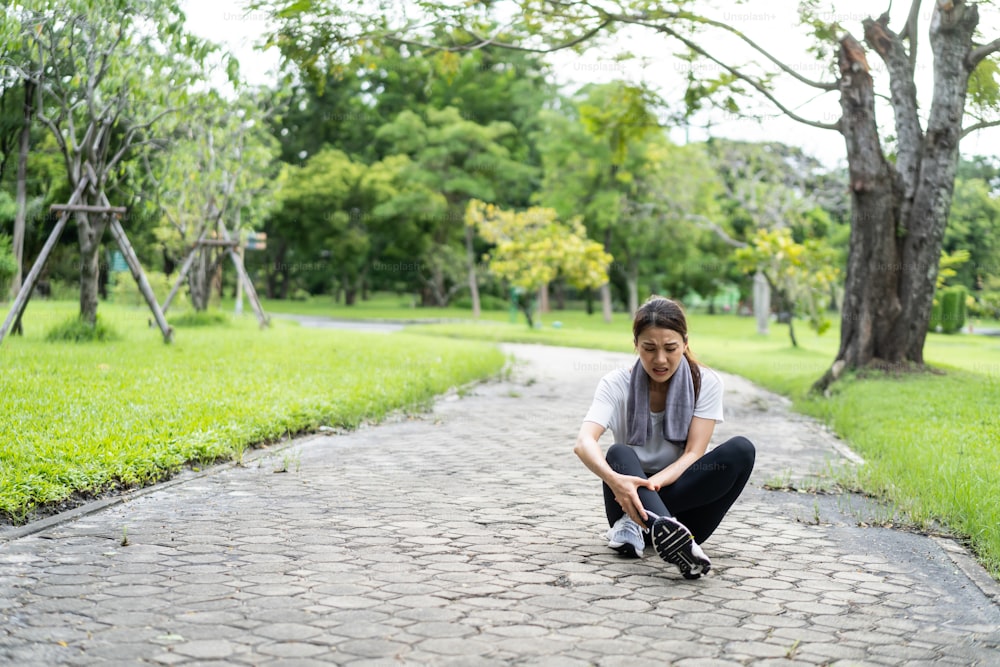 Une femme sportive asiatique attrayante ressent une douleur à la cheville lorsqu’elle court dans la rue. Belle athlète en tenue de sport massant et étirant ses jambes d’un problème de ligament articulaire après avoir fait de l’exercice dans un parc public.