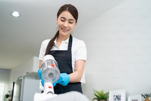 Asian cleaning service woman worker cleaning in living room at home. Beautiful happy girl housewife housekeeper cleaner wear apron and vacuuming messy dirty table for housekeeping housework or chore.