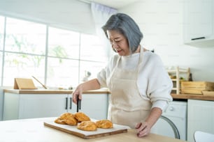 Portrait of Asian happy Senior elderly woman stand in kitchen at home. Attractive Older grandmother feeling happy and enjoy retirement life, cooking foods bake bakery and smiling in house.