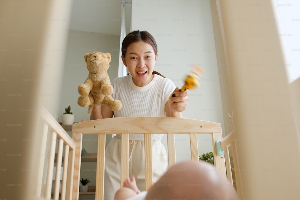 Young Asian mother playing doll with her newborn baby on crib joyfully