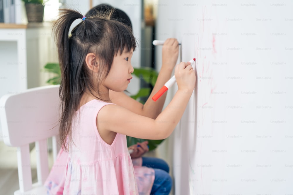 아시아 어린 형제 소녀가 거실의 흰 벽에 페인트를 칠하는 것을 즐긴다. 행복으로 예술 그림을 그리며 색칠하는 재미를 느끼는 작고 사랑스러운 아이들은 집에서 휴가를 보내는 창의력 활동을 즐깁니다.