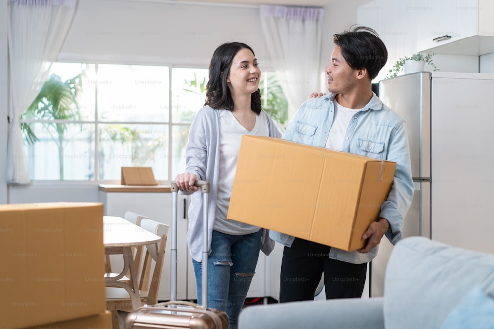 Asian young happy new marriage couple moving to new house together. Attractive romantic man and woman holding box parcel and suitcase with happiness and love. Family-Moving house relocation concept.