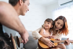 집에서 어린 아이와 함께 기타를 연주하는 아시아 젊은 부부. 매력적인 아름다운 부모님은 소파에 앉아 집안의 거실에서 노래를 부르는 어린 딸과 여가 시간�을 보냅니다.
