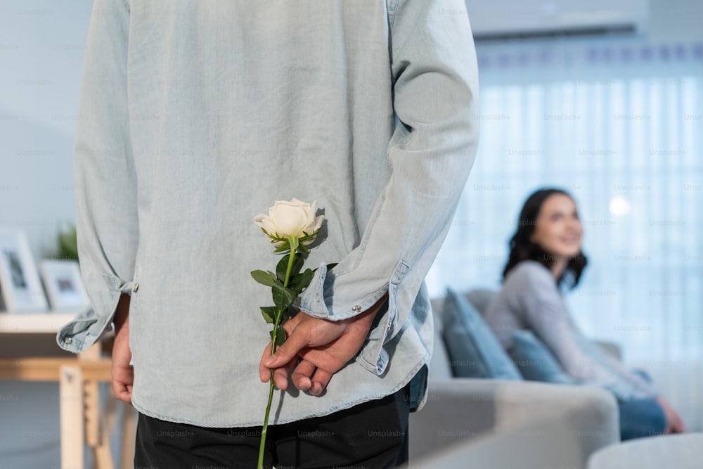 Asiatischer junger Mann Überraschung und geben Rosenblume zu schönen Freundin. Attraktives romantisches neues Ehepaar Mann und Frau verbringen Zeit miteinander im Wohnzimmer zu Hause, feiern Jubiläum im Haus
