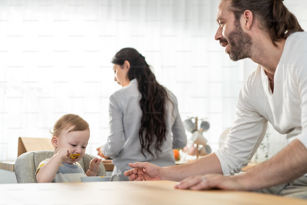 Caucasian beautiful parents take care of baby boy toddler in kitchen. Happy family, Attractive young couple cook and feed healthy foods to little kid son infant while sit on chair for lunch in house.