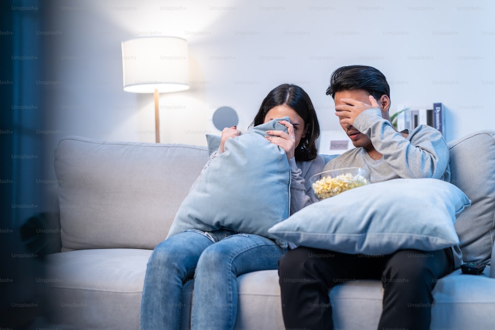 Jovem casal asiático engraçado assistir ao filme jump scare na televisão em casa. Jovem novo casamento homem e mulher se sentem animados e chocados enquanto se sentam no sofá e assistem a vídeos assustadores na TV na sala de estar em casa.