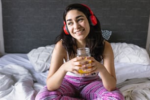 メキシコラテンアメリカの自宅のベッドでヘッドフォンで音楽を聴き、オレンジジュースを飲む若いラテン系女性