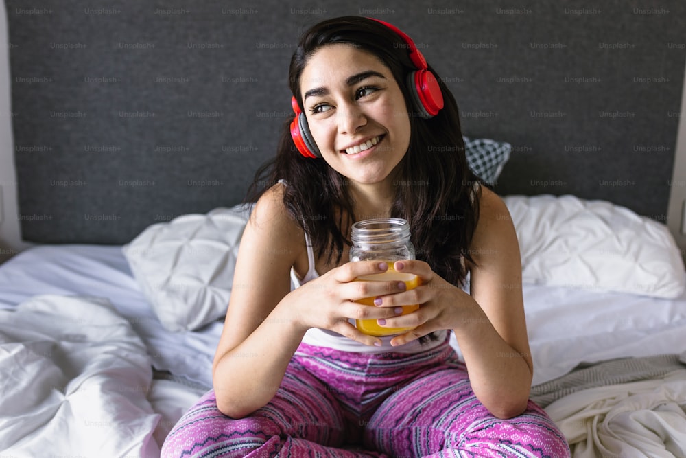 Young latin woman listening music with headphones and drinking orange juice on bed at home in Mexico Latin America