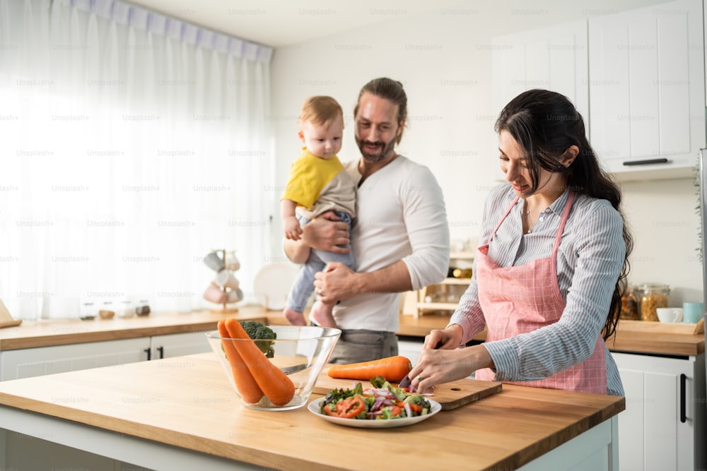 Caucasian beautiful parents cook food with baby boy toddler in kitchen. Happy family, Attractive young mother making healthy salad for lunch while husband father holding little kid son infant in house