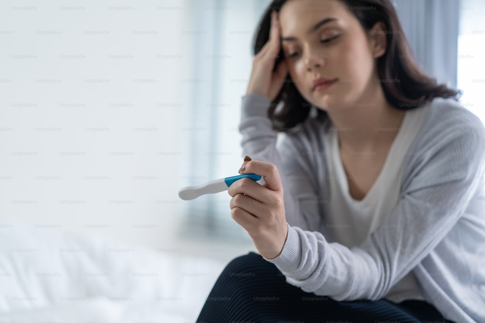 Asian young woman feeling shock after look at positive Pregnancy test. Disappointed attractive beautiful unplanned pregnancy girl sit on bed with upset and sad face for problem don't want to pregnant.