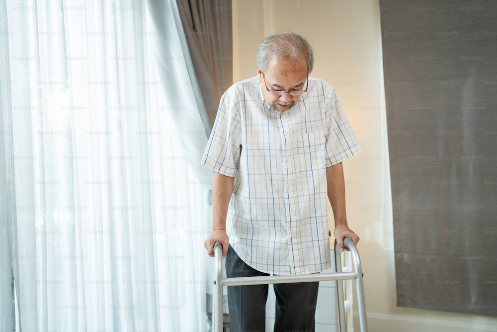 Asian Senior elderly man patient walking slowly with walker in house. Mature older grandfather doing physical therapy alone with cane in living room at home. Medical Health care insurance concept.