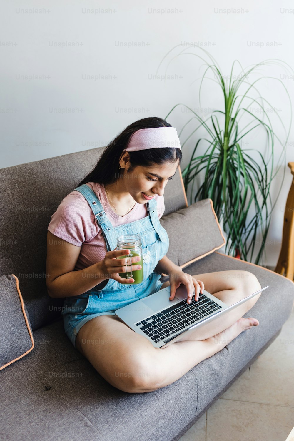 young latin Woman student using laptop in a video call or online class and drinking green juice while sitting on sofa at home in Mexico