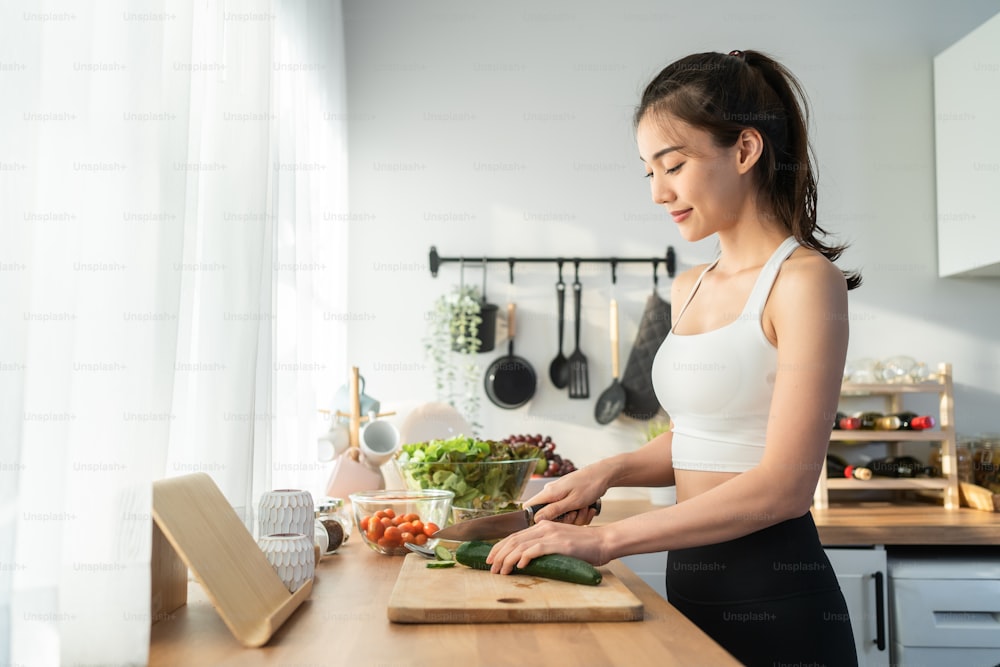 Asaian attractive woman wear sport clothes cook green salad in kitchen. Young beautiful girl feeling happy and enjoy eating vegetables healthy foods to diet and lose weight for health care in house.