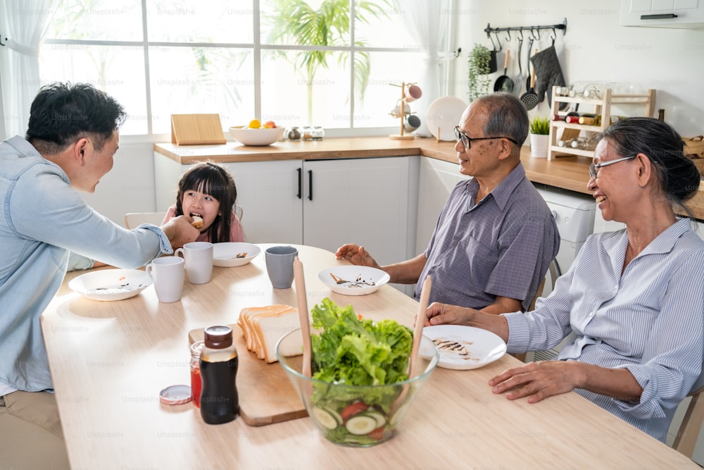 La gran familia asiática feliz almuerza en la mesa de comer juntos en la casa. El abuelo mayor, el padre joven y la hija pequeña se sienten felices, disfrutan comiendo pan en casa. Concepto de relación de actividad.