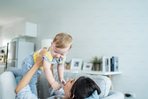 Caucasian loving mom play with baby boy child on sofa in living room. Happy family, attractive beautiful caring young mother lifting toddler son fly up enjoy doing airplane game in morning in house.