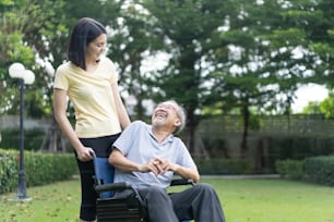 Asian happy elder retired grandfather spend time outdoor at green park. Attractive girl daughter push senior father on wheelchair with smile face, enjoy hearth care activity. Medical Insurance concept