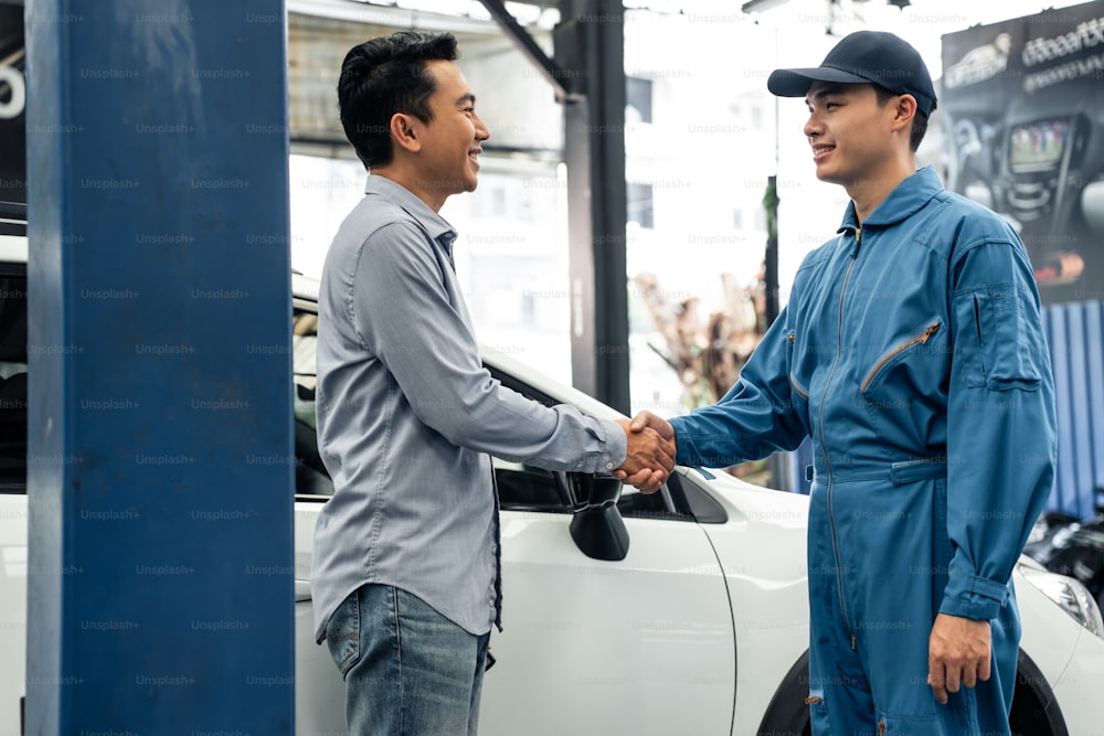ガレージでクライアントと握手するアジアの自動車整備士の修理工。メカニックワークショップで働く車両サービスマネージャーは、顧客の車のエンジンをチェックし、メンテナンスした後、幸せと成功を感じます。