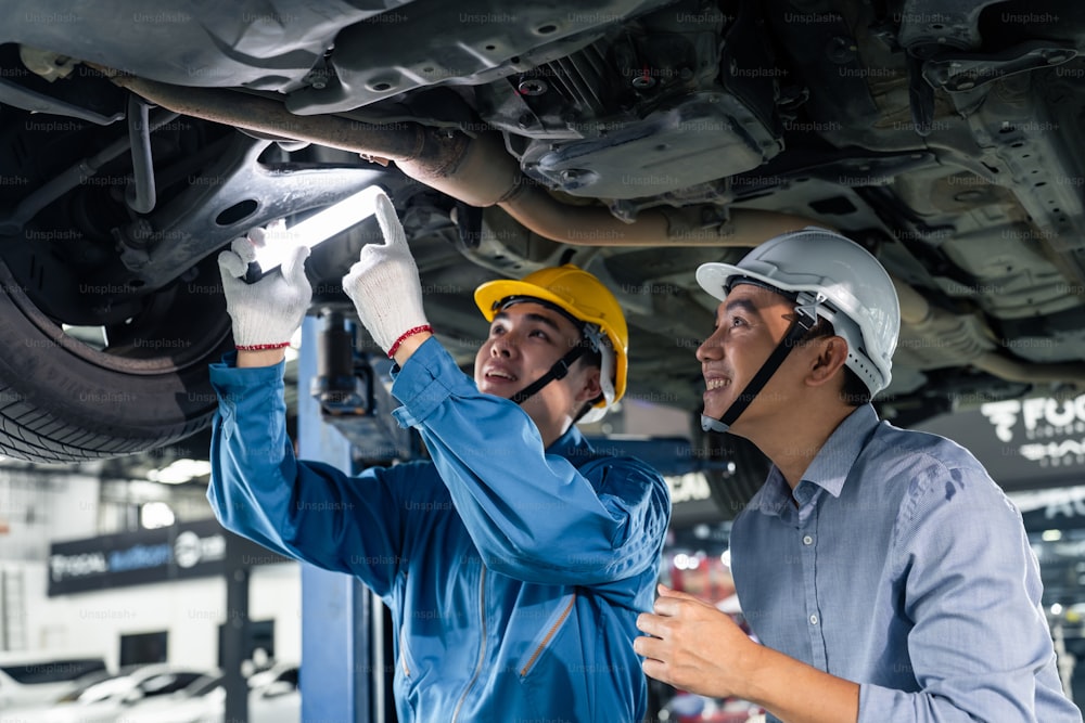 アジアの自動車整備士がガレージでクライアントに車の状態を説明します。整備工場での車両サービスマネージャーの男性の仕事は、メンテナンスと修理車のエンジンのために顧客の男性に車両の部品を指しています