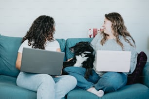 two women sitting on a couch with laptops and a dog