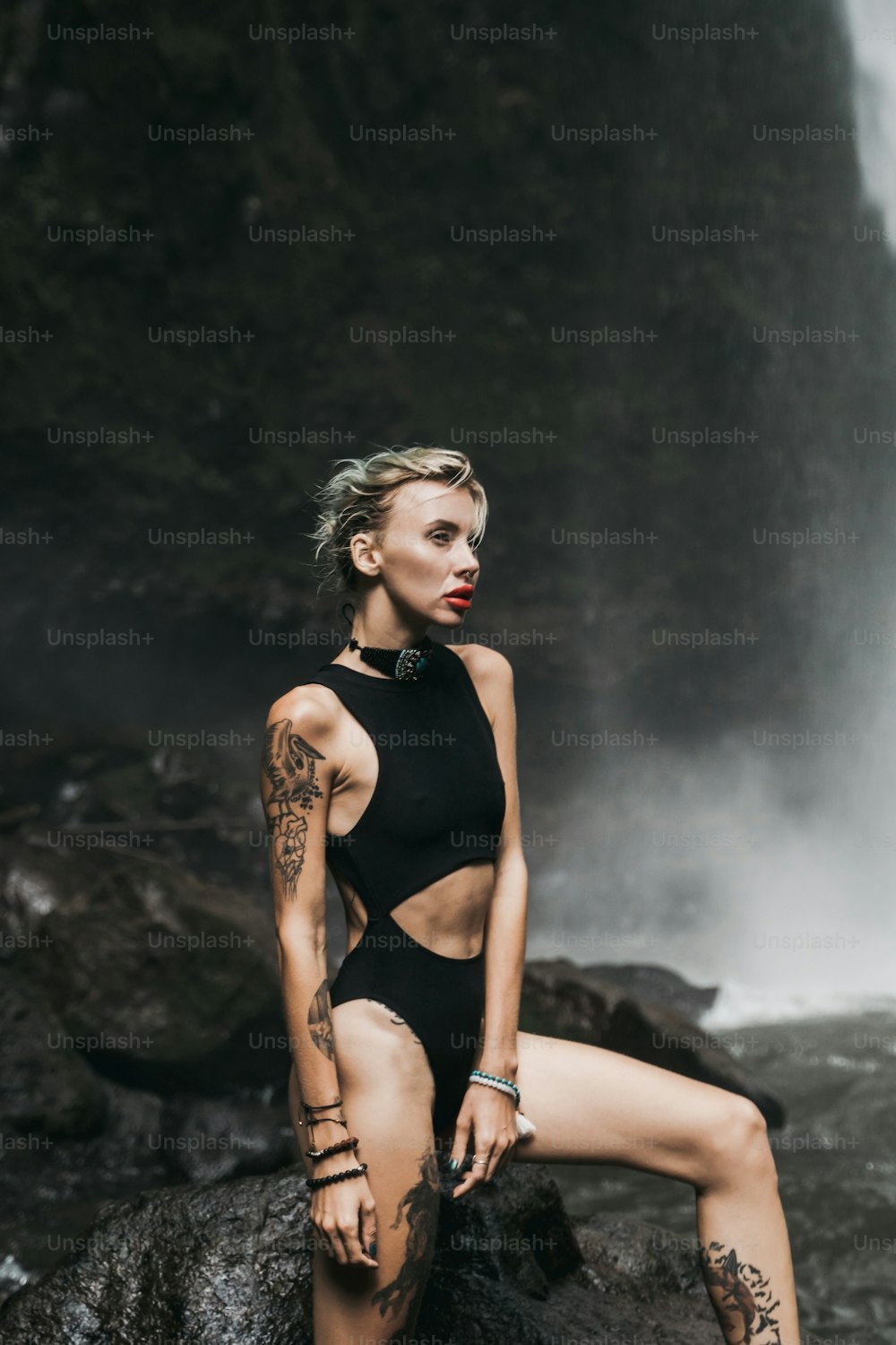 a woman in a black swimsuit sitting on a rock near a waterfall