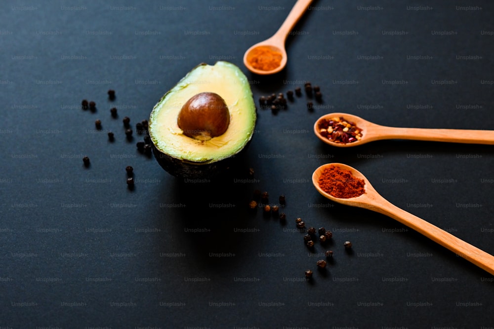an avocado and spoons of spices on a black surface