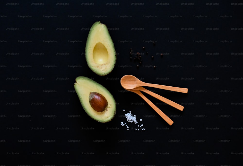 an avocado, spoons, salt and pepper on a black surface