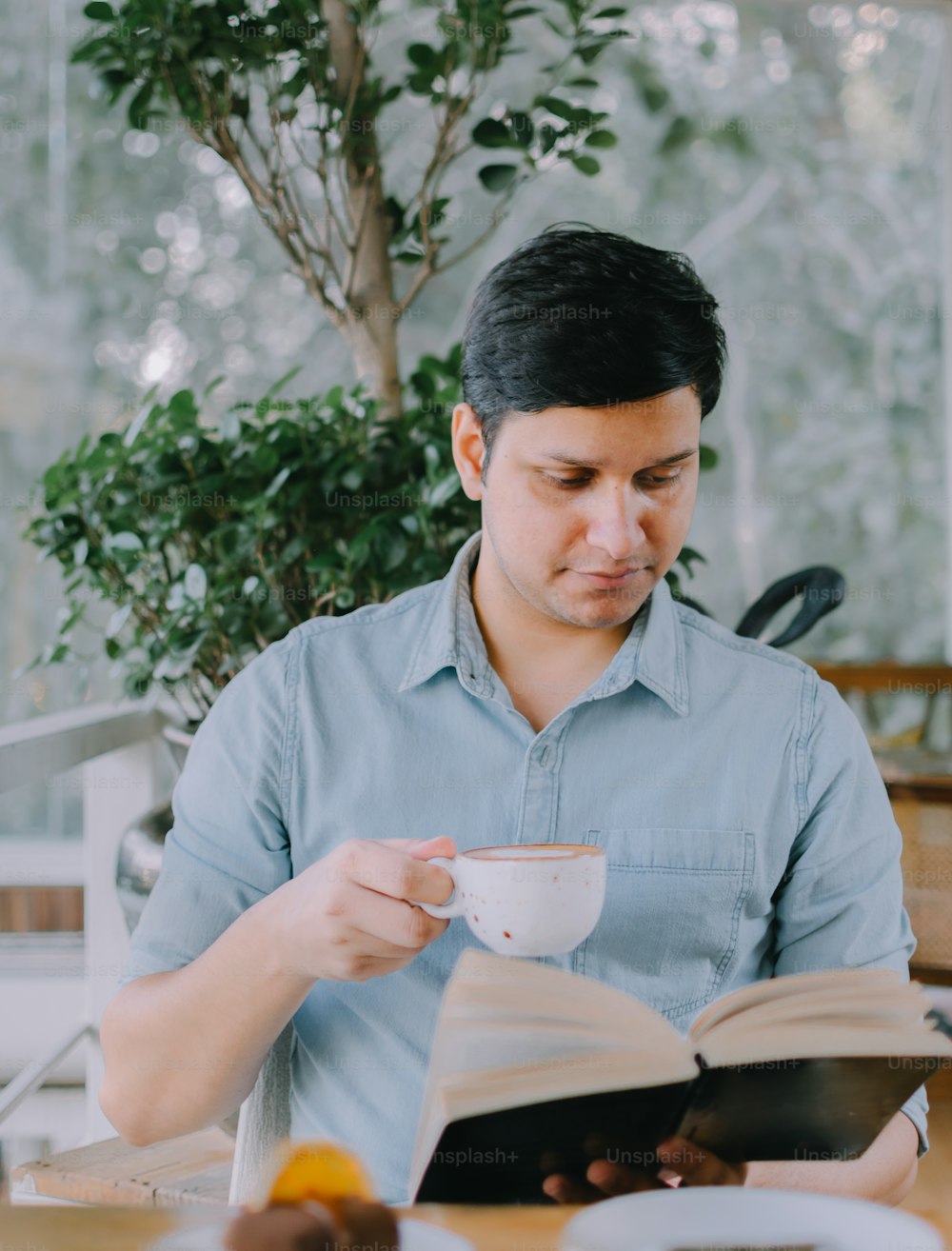 a man sitting at a table reading a book and drinking a cup of coffee